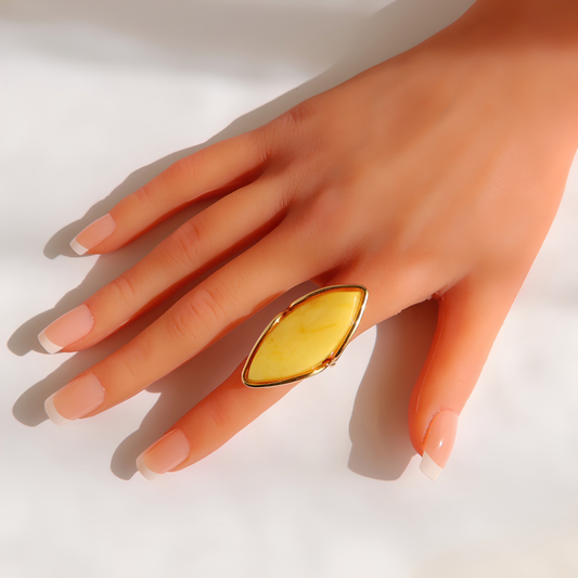 A close-up of a hand wearing a large gold butterscotch amber ring with a marquise-shaped yellow stone, set against a bright white background with soft shadows.