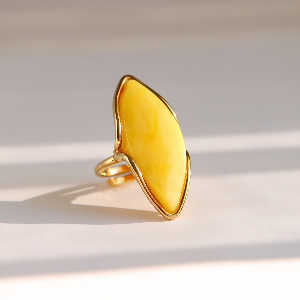 A gold butterscotch amber ring featuring a large, organic-shaped yellow stone, photographed on a light beige surface with soft diagonal shadows.