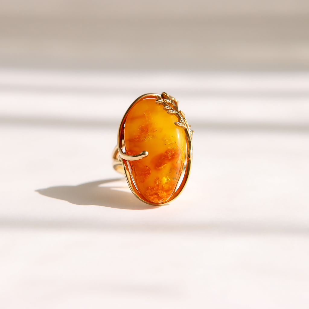 An oval amber ring with a gold leaf-wrapped setting, photographed on a warm off-white surface with soft shadows.