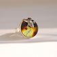 A silver amber ring featuring a round cabochon stone with a marbled pattern of dark brown, amber, and yellow-green, adorned with a textured silver frog  photographed on a clean white background.
