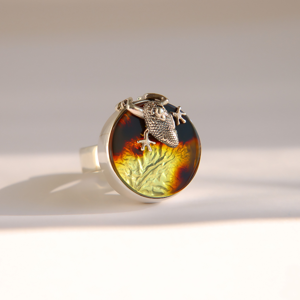 A silver amber ring featuring a round cabochon stone with a marbled pattern of dark brown, amber, and yellow-green, adorned with a textured silver frog  photographed on a clean white background.