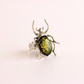 A silver green amber beetle ring featuring a large, polished olive-green oval gemstone in the center, set against a solid pale pink background.