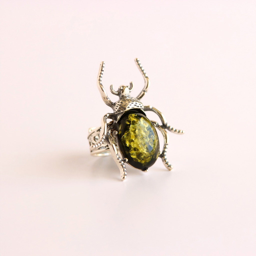 A silver green amber beetle ring featuring a large, polished olive-green oval gemstone in the center, set against a solid pale pink background.