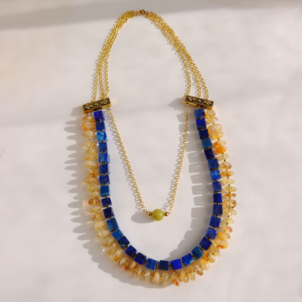A gold-tone multi-strand statement necklace featuring blue and amber-orange stone beads, displayed on a light off-white background.
