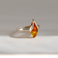 A silver amber ring featuring a teardrop-shaped amber gemstone with rich orange and yellow hues, set against a clean, light gray background.