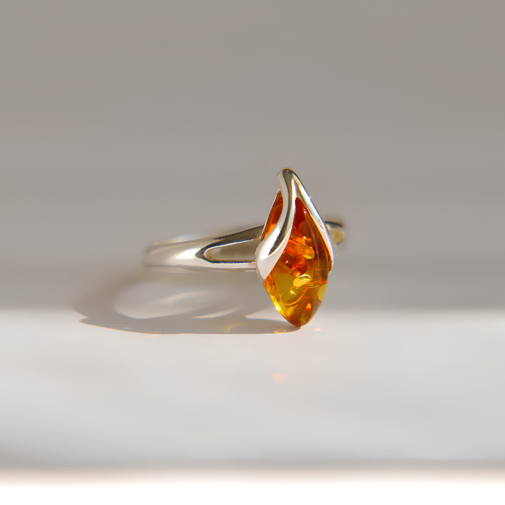 A silver amber ring featuring a teardrop-shaped amber gemstone with rich orange and yellow hues, set against a clean, light gray background.