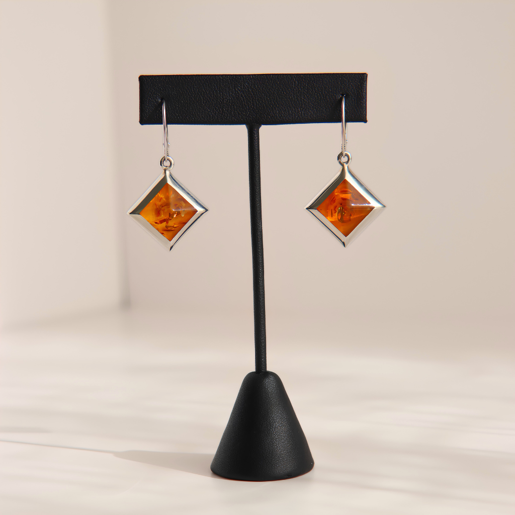 A pair of silver drop amber earrings with diamond-shaped amber inlays, displayed on a black jewelry stand against a warm off-white background.