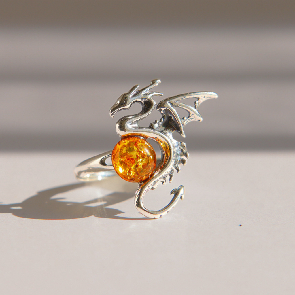 A silver amber dragon ring with a round, glittering amber-orange gemstone, photographed on a neutral light gray surface with soft shadows.