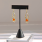A pair of gold spider-shaped dangle amber earrings with round orange gemstones, displayed on a black jewelry stand against a soft gray background.