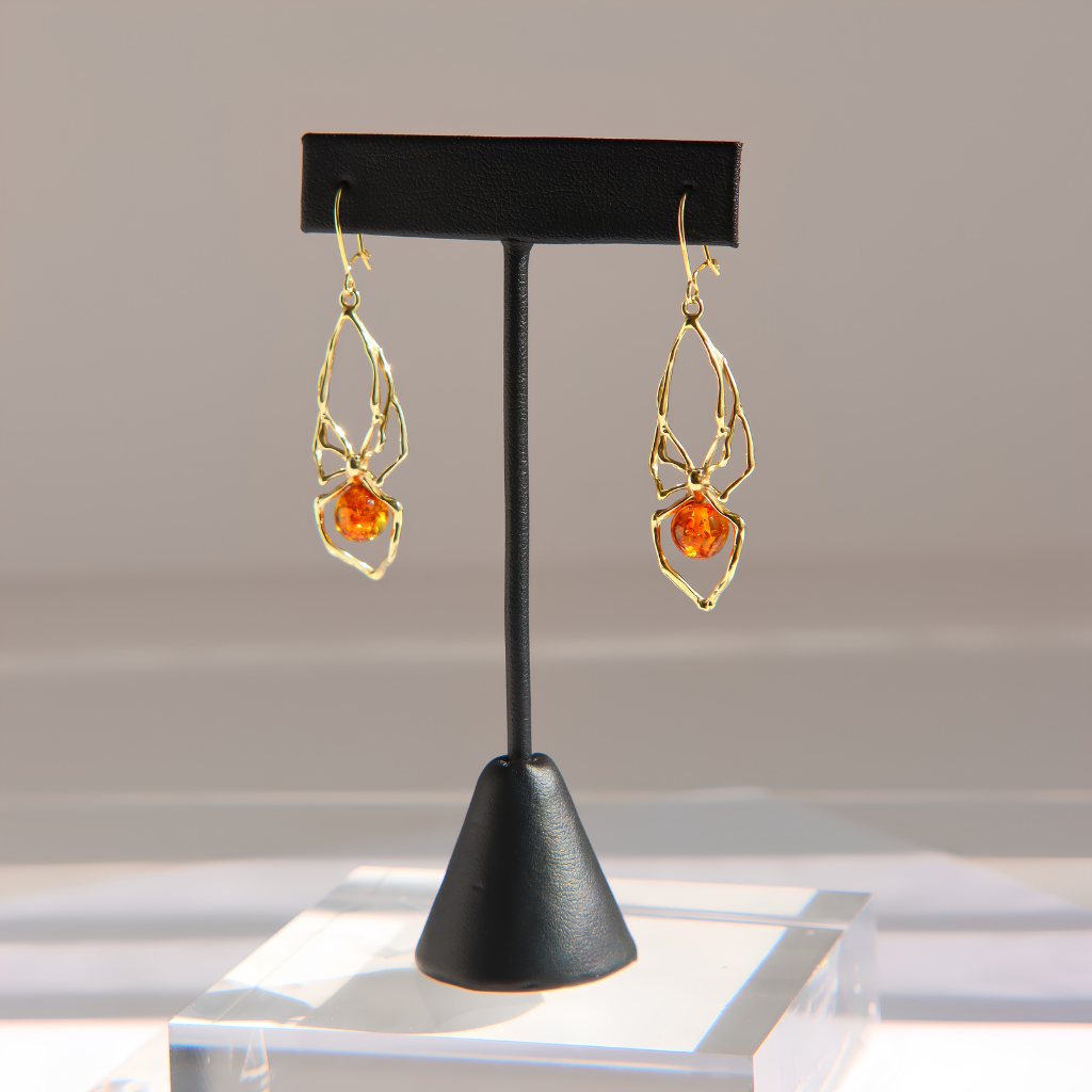 A pair of gold spider-shaped dangle amber earrings with round orange gemstones, displayed on a black jewelry stand against a soft gray background.