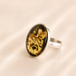 A silver amber cameo ring featuring an oval black cabochon stone with intricate gold floral and butterfly engravings, set against a soft beige satin background.
