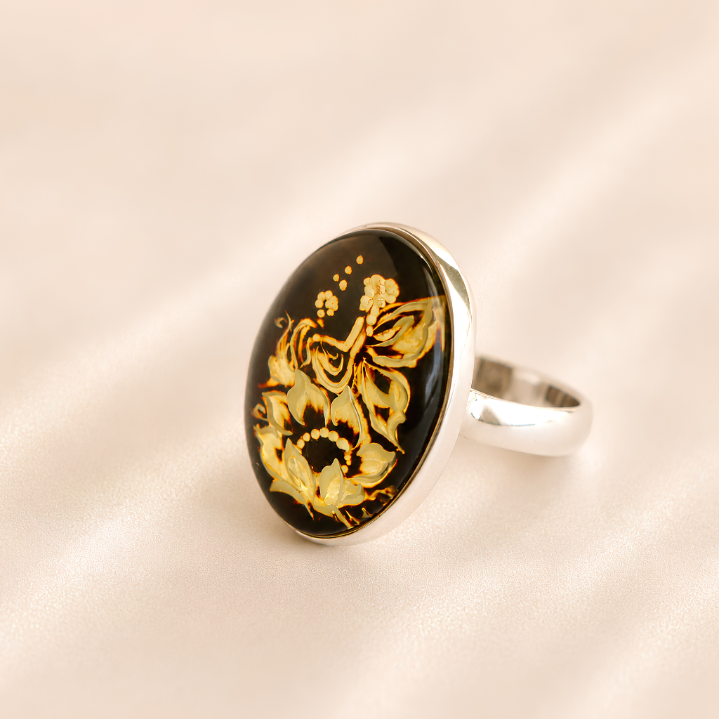 A silver amber cameo ring featuring an oval black cabochon stone with intricate gold floral and butterfly engravings, set against a soft beige satin background.