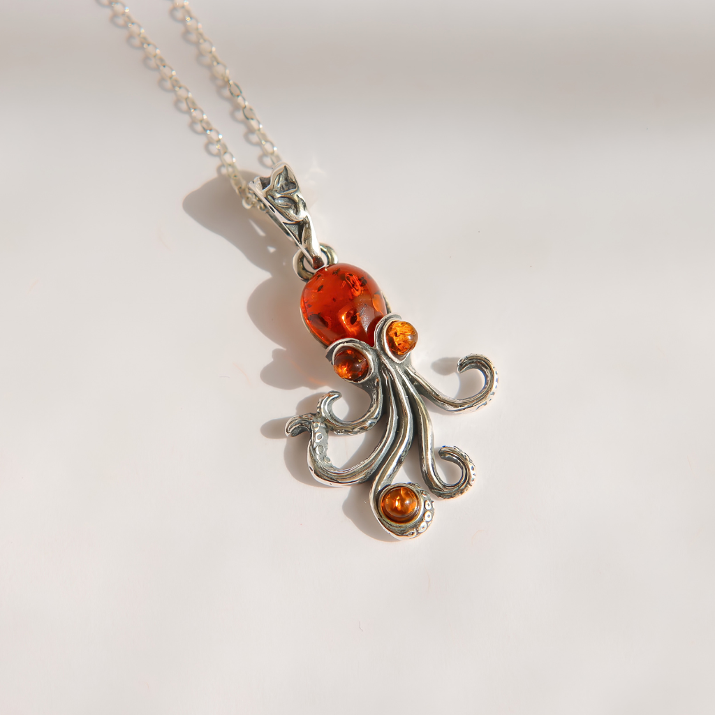 A silver pendant amber necklace featuring a stylized octopus design with a large round amber-orange cabachon on the head and three smaller amber-orange beads on the tentacles, shown on a white background.