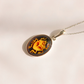 An oval pendant amber scarab necklace featuring a yellow and orange beetle motif on a dark background, suspended from a silver chain against a warm off-white background.