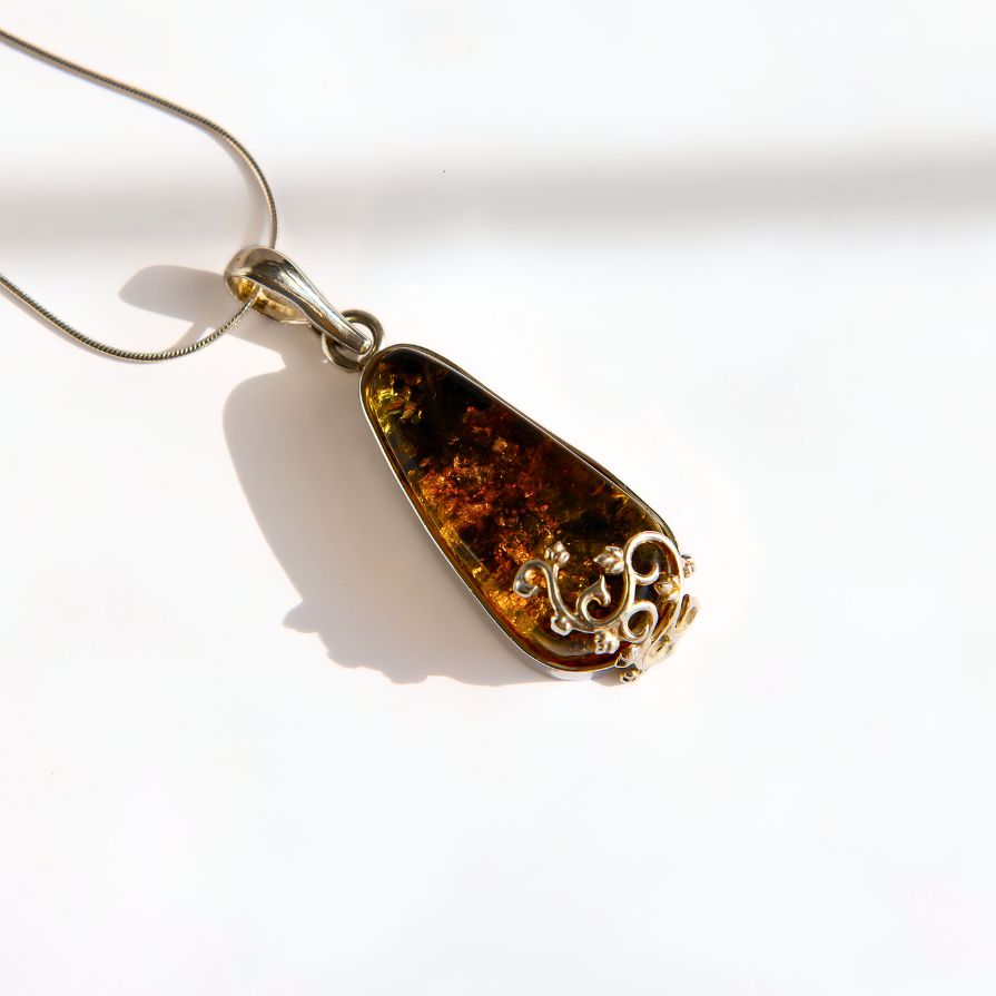 A teardrop-shaped green amber pendant necklace with a silver-toned filigree accent on one end, hanging from a silver chain against a bright white background.