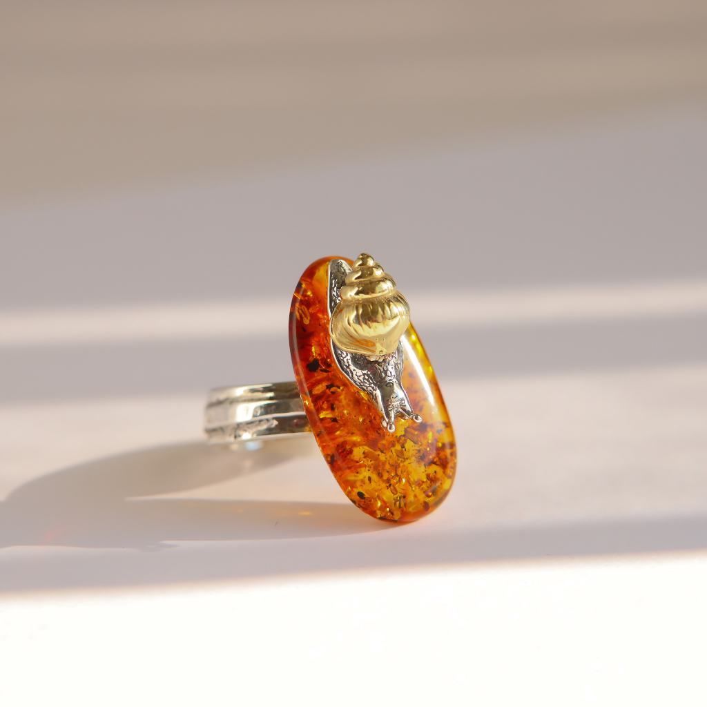 A silver amber ring featuring a large, teardrop-shaped amber cabochon with a detailed gold snail mounted on top, set against a warm, sunlit beige background.