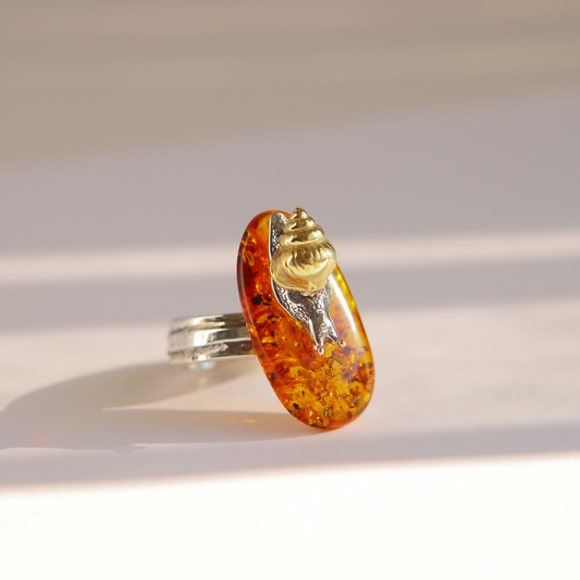 A silver amber ring featuring a large, teardrop-shaped amber cabochon with a detailed gold snail mounted on top, set against a warm, sunlit beige background.