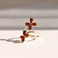 A Baltic amber flower ring in sterling silver featuring two small four-petal flower motifs set with polished amber-red stones, photographed on a light beige surface with soft shadows.