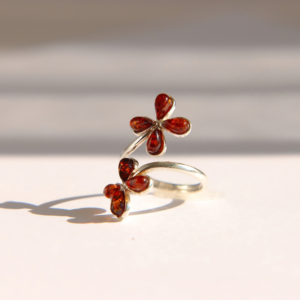 A Baltic amber flower ring in sterling silver featuring two small four-petal flower motifs set with polished amber-red stones, photographed on a light beige surface with soft shadows.
