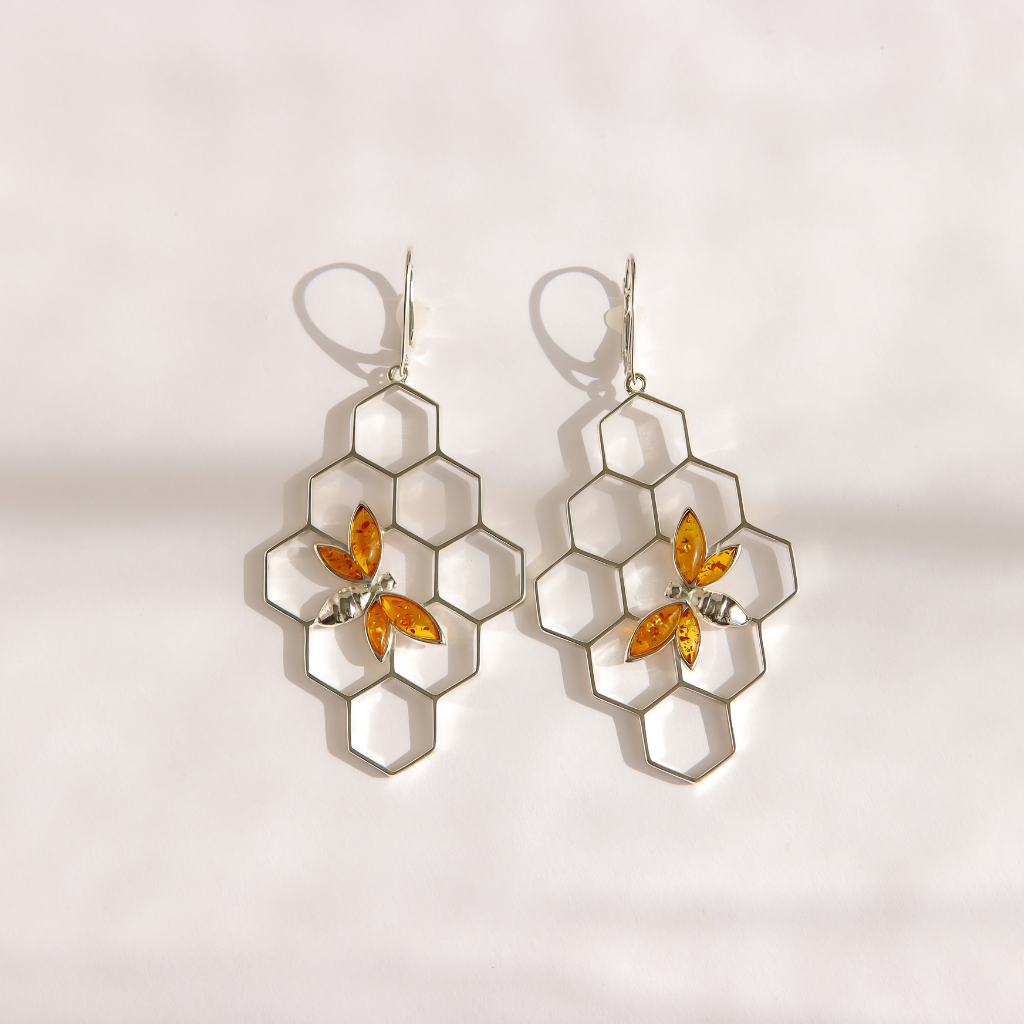 A pair of silver-toned dangle bee earrings featuring a honeycomb pattern with amber-colored bee accents, displayed on a light, textured background.
