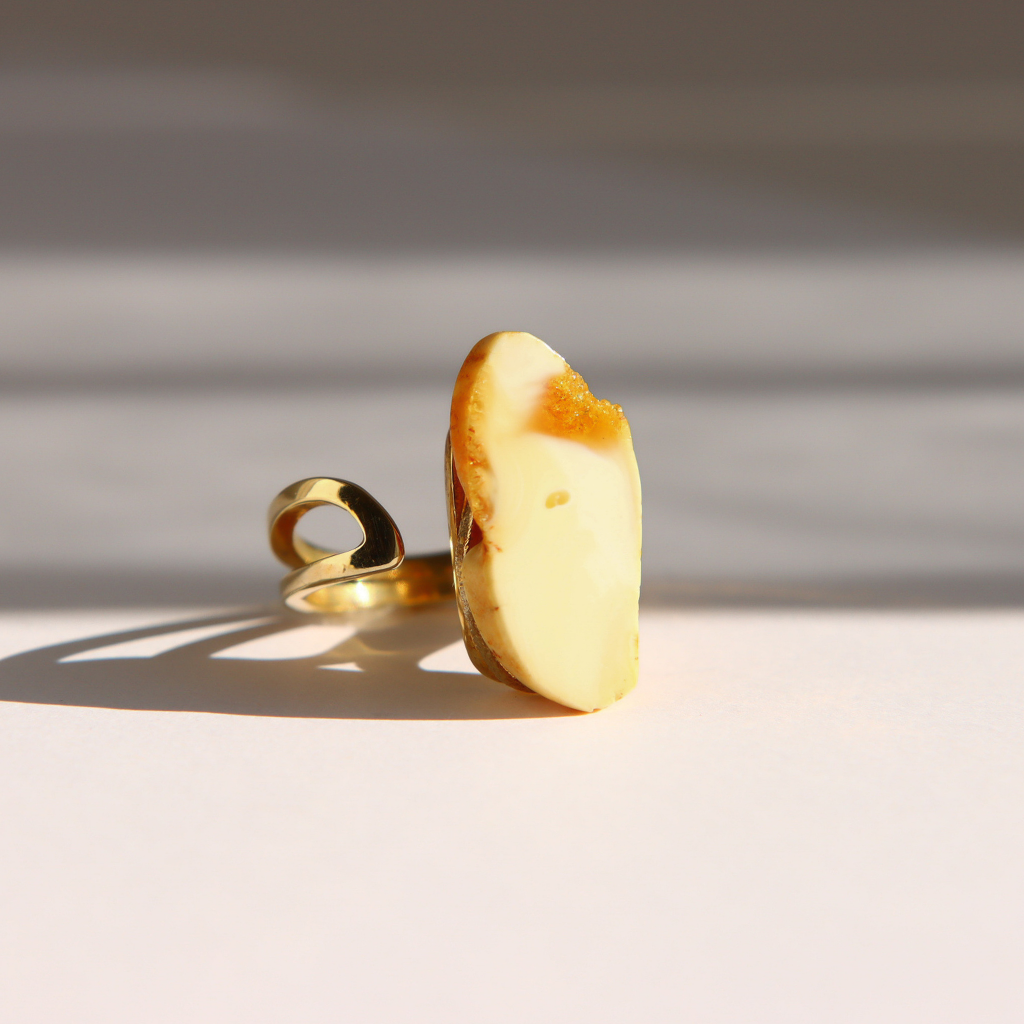 A gold white amber ring with a large, irregularly shaped cream-colored stone, standing upright on a light surface with dramatic diagonal shadows.