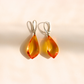 A pair of teardrop-shaped Baltic amber drop earrings with silver leverback hooks, displayed on a warm beige background with soft shadows.