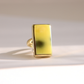 A gold Baltic amber ring featuring a large, rectangular yellow stone with a dark, cloudy center, photographed on a light beige background with soft shadows.