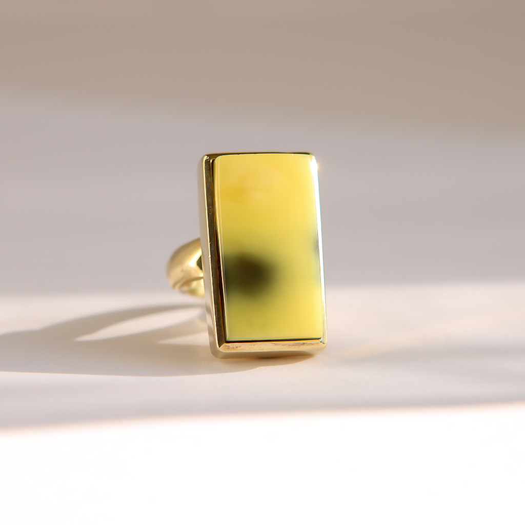 A gold Baltic amber ring featuring a large, rectangular yellow stone with a dark, cloudy center, photographed on a light beige background with soft shadows.