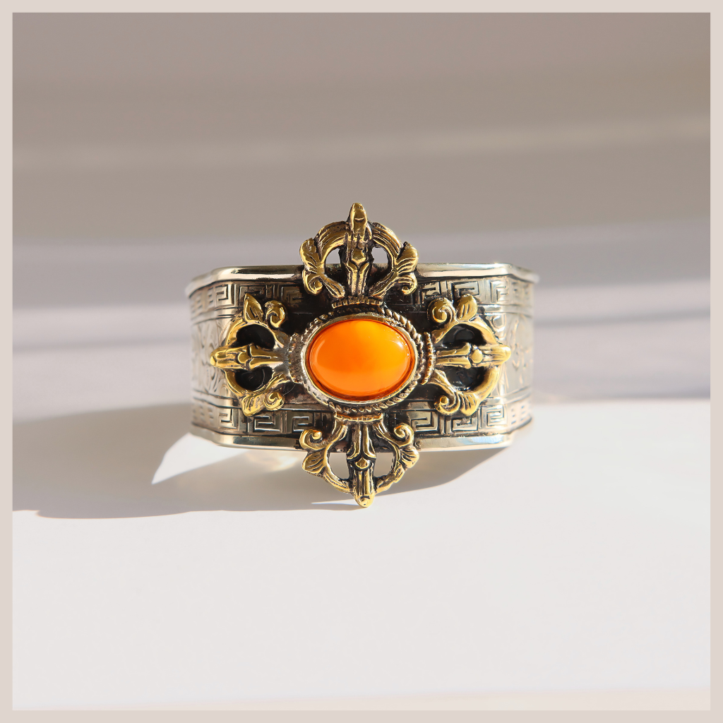A wide silver-toned amber cuff bracelet with ornate gold-toned filigree details and a central oval orange stone, photographed on a light neutral background.