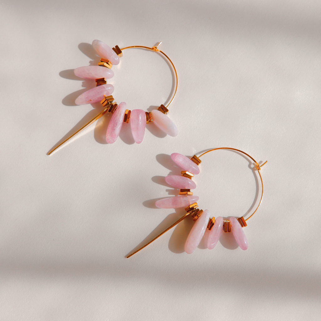 A pair of gold kunzite hoop earrings with dangling pink stone beads and small gold spacer rings, photographed on a warm off-white background with soft shadows.