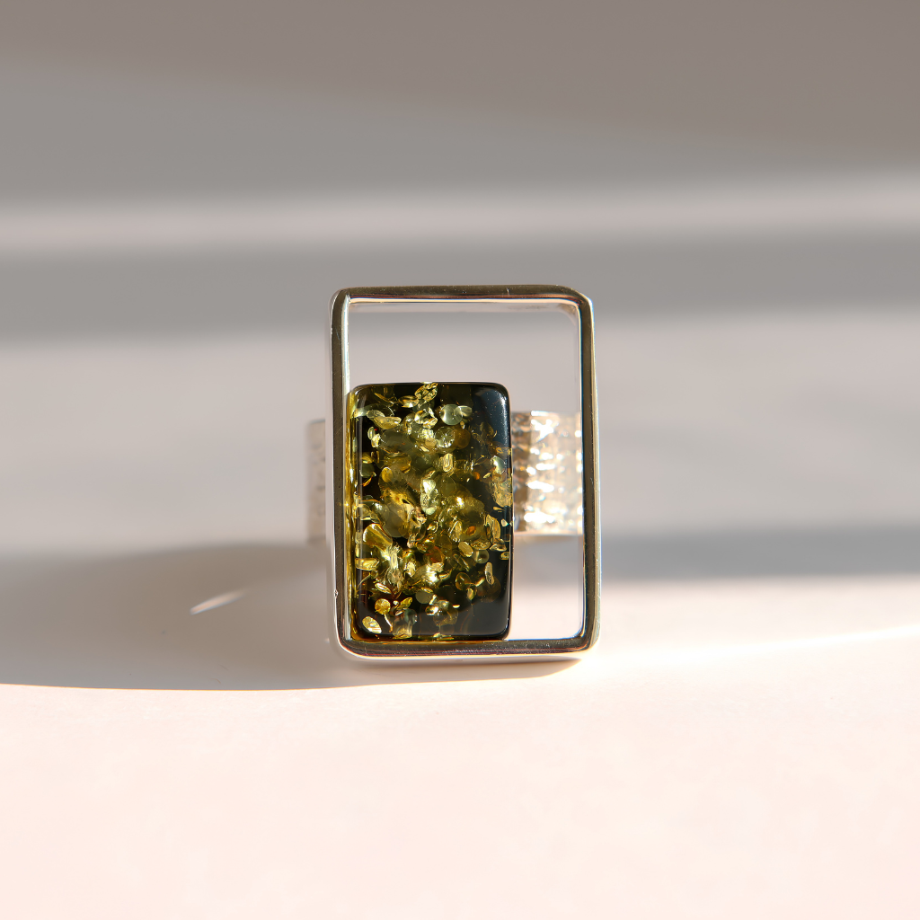 A rectangular silver amber ring featuring a central bezel-set piece with a dark black center and a textured olive-green outer layer, photographed on a light beige surface with soft diagonal shadows.
