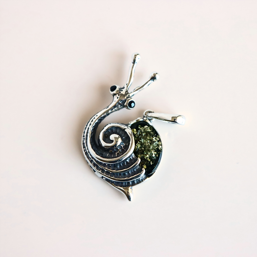 A silver amber snail pendant with a textured spiral shell, featuring a green amber inlay and small black bead eyes, shown on a plain light pink background.