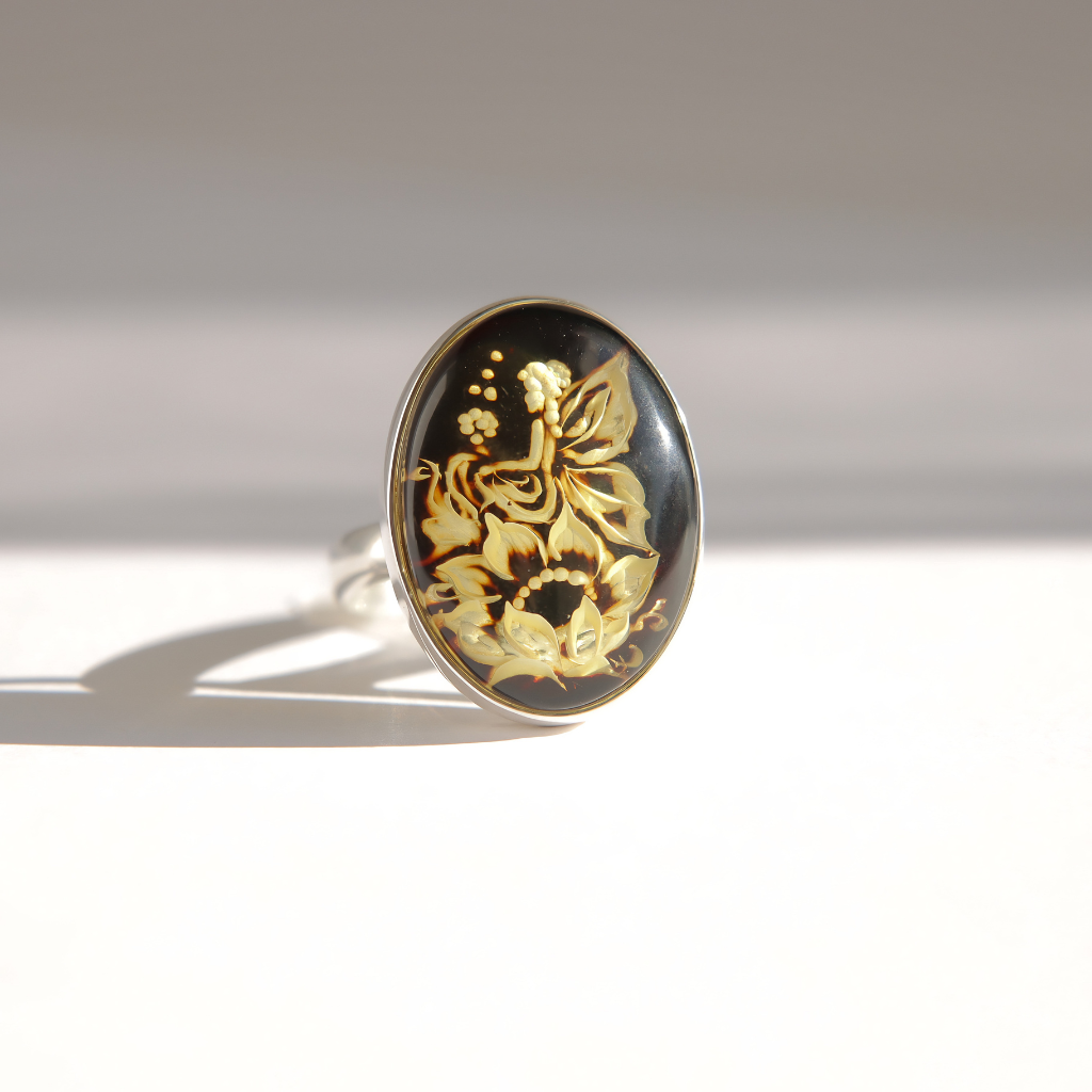 A silver amber cameo ring with an oval black amber cabochon featuring a carved fairy and floral design, photographed on a white surface with a soft shadow.