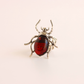 A scarab ring featuring a polished dark red oval cabochon stone set in silver-toned metal with detailed legs and antennae, shown on a plain light background.
