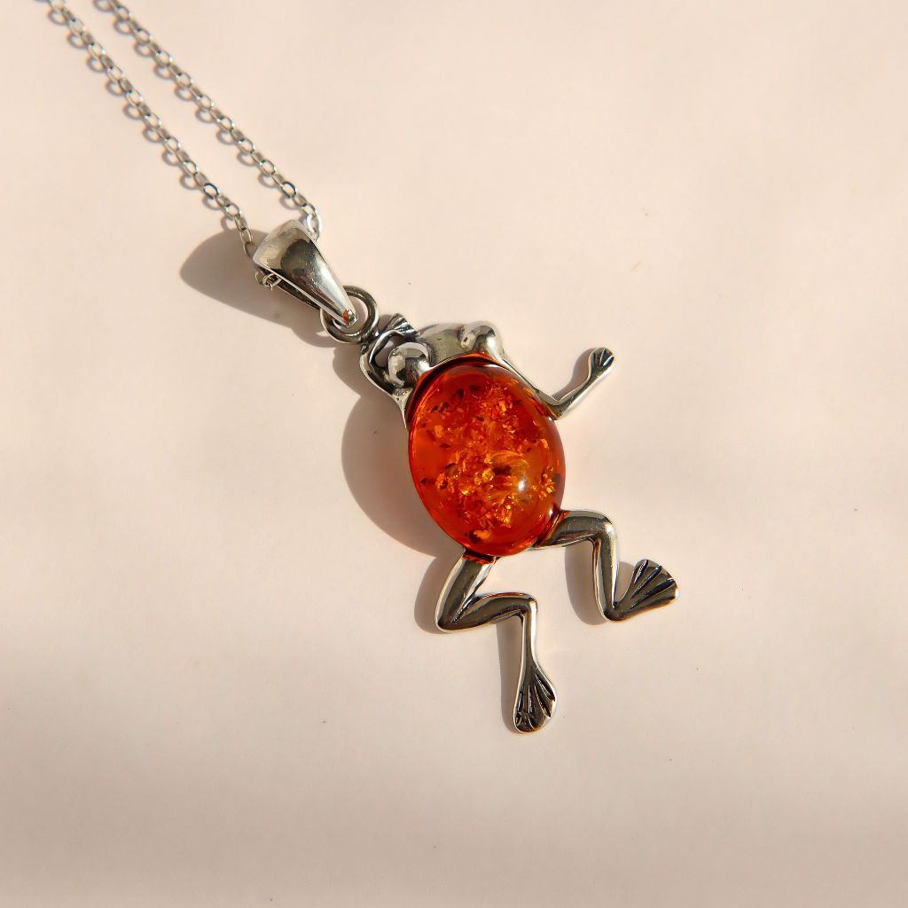 A silver amber frog pendant necklace with a vibrant orange-red amber glass body, shown on a light beige background.