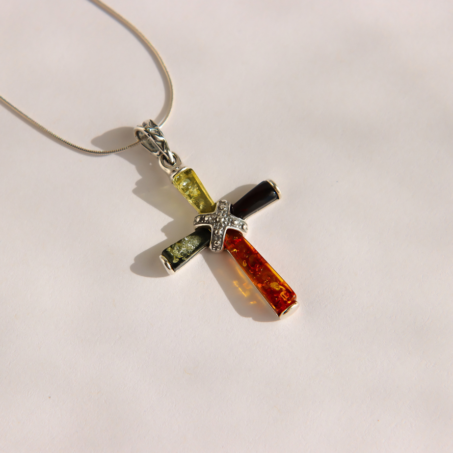 A silver amber cross pendant necklace featuring four rectangular amber segments in yellow-green, black, orange, and green, with a sparkling silver center, on a thin silver chain against a light background.