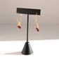 A pair of gold hoop earrings with small dangling red gemstone drops, displayed on a black jewelry stand against a light gray background.