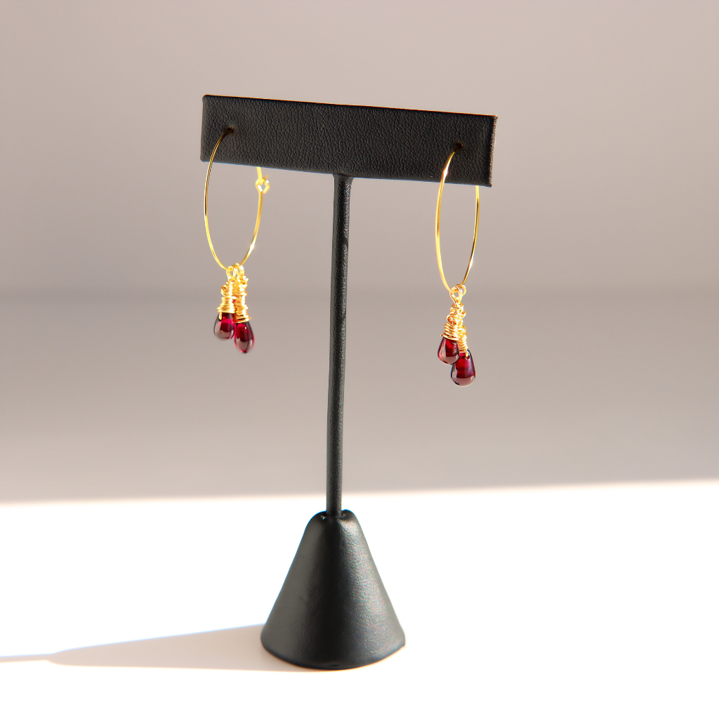 A pair of gold hoop earrings with small dangling red gemstone drops, displayed on a black jewelry stand against a light gray background.