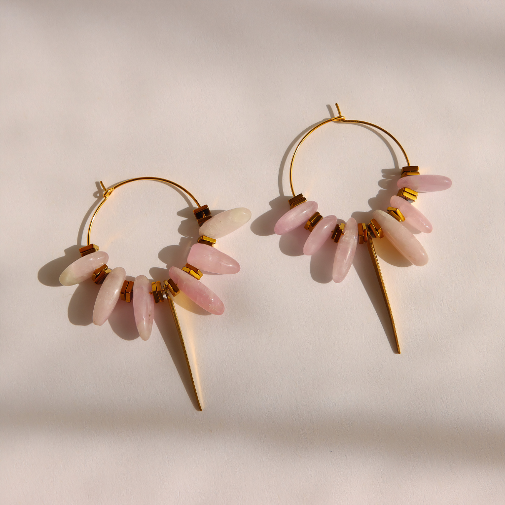 A pair of gold kunzite dagger hoop earrings with dangling pale pink stone beads and cream-colored cone charms, displayed on a warm off-white background with soft shadows.