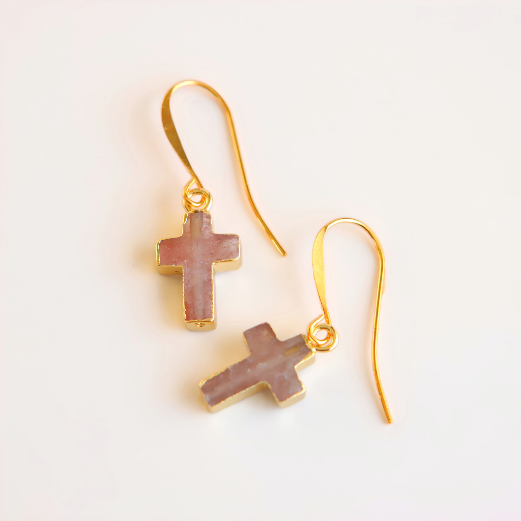 A pair of gold fishhook drop earrings with small rose quartz cross charms featuring a dusty pink textured inlay, set against a plain white background.