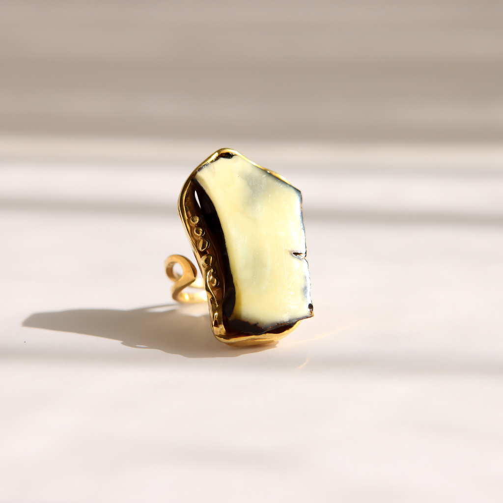 A gold amber statement ring with a large, irregularly shaped cream-colored stone, photographed on a light beige surface with soft shadows.