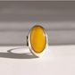 An oval sterling silver amber poison ring with a polished yellow amber cabochon stone set in a silver bezel, standing upright on a light gray surface with soft shadows.