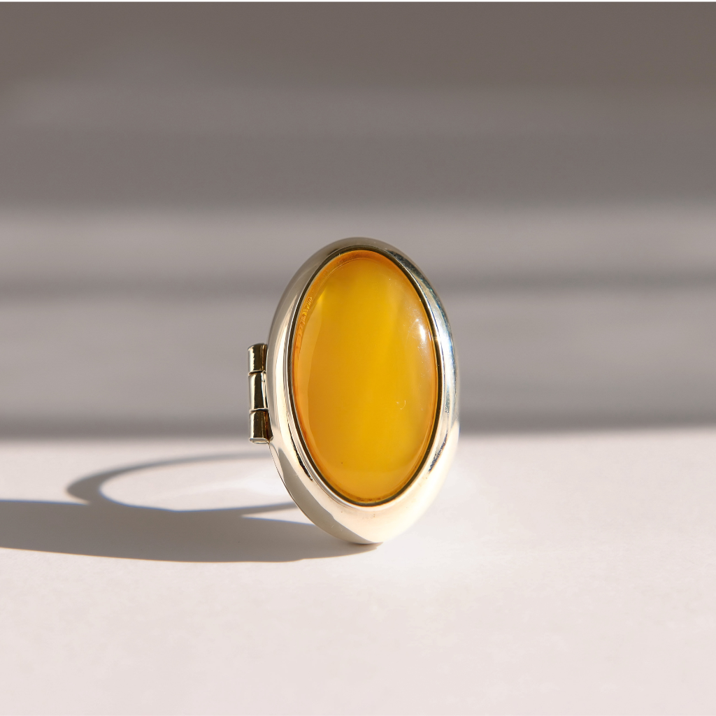An oval sterling silver amber poison ring with a polished yellow amber cabochon stone set in a silver bezel, standing upright on a light gray surface with soft shadows.