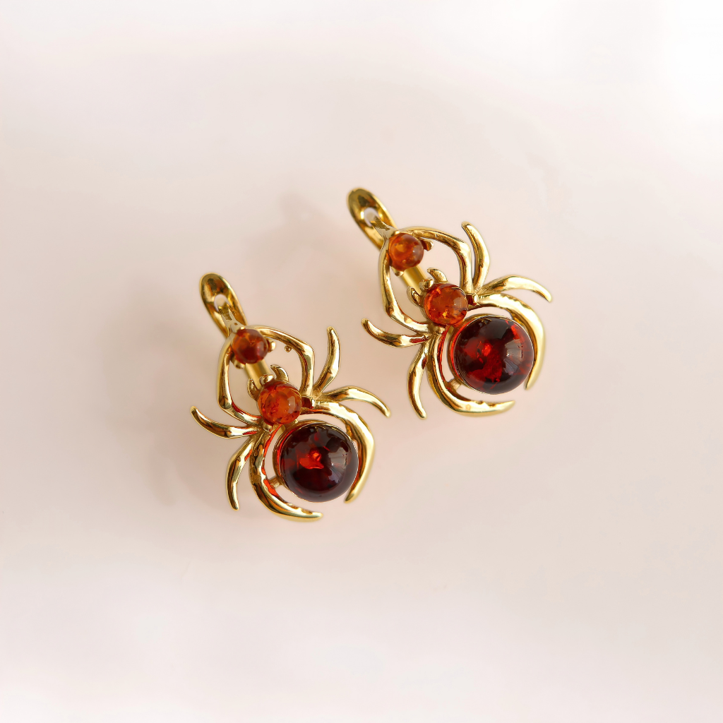 A pair of gold spider-shaped earrings with faceted dark red gemstones and small amber-orange accent stones, shown on a clean white background.
