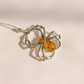 A amber spider-shaped pendant necklace made of silver-toned metal with a large amber-colored oval center and a smaller round orange accent, shown on a plain light background.