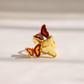 A gold-plated amber butterfly ring with cream, red, and orange amber details, resting on a light beige surface with soft shadows.