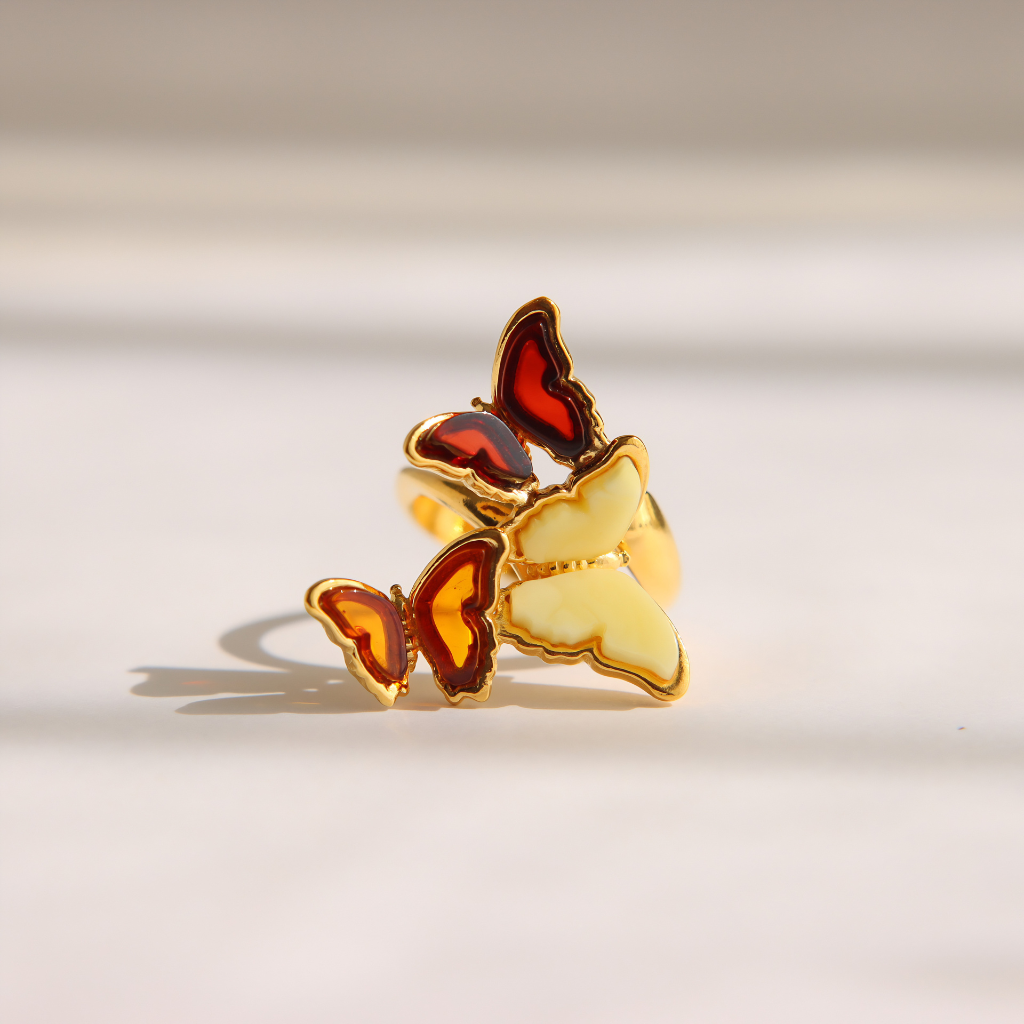 A gold-plated amber butterfly ring with cream, red, and orange amber details, resting on a light beige surface with soft shadows.