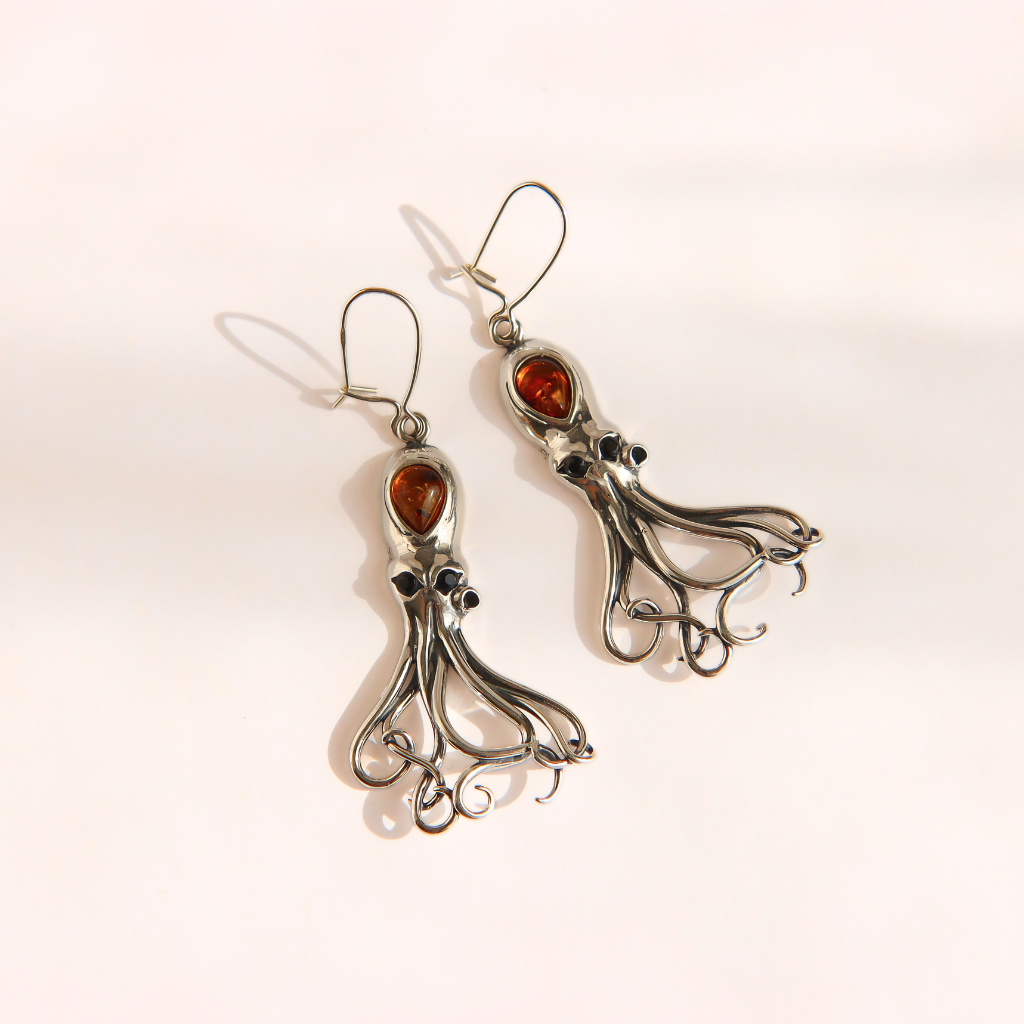 A pair of silver-toned amber octopus dangle earrings with amber-brown gemstone eyes and small black bead accents, photographed on a clean white background.