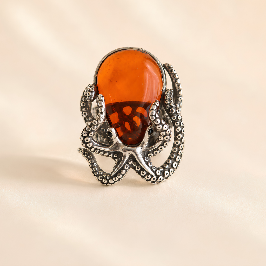 A silver octopus ring featuring a large, smooth, rounded amber-orange cabochon gemstone on its back, set against a soft beige background.