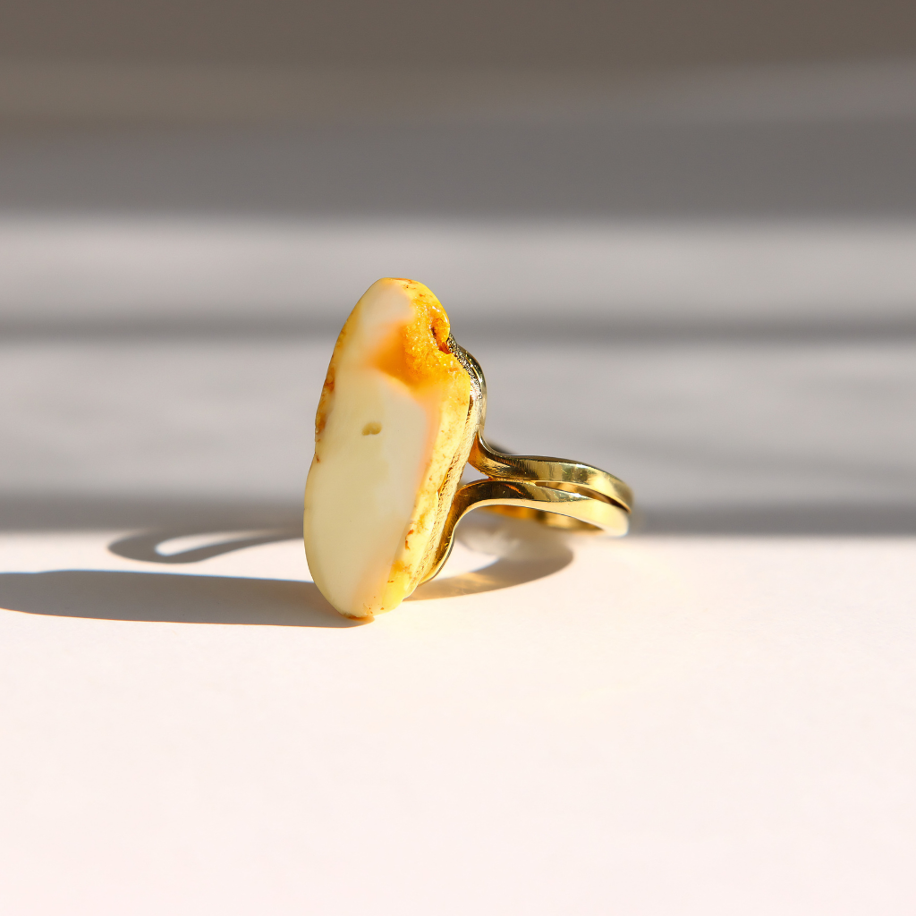A gold white amber ring with a large, irregularly shaped cream-colored stone featuring orange-brown edges, photographed on a white surface with soft diagonal shadows.
